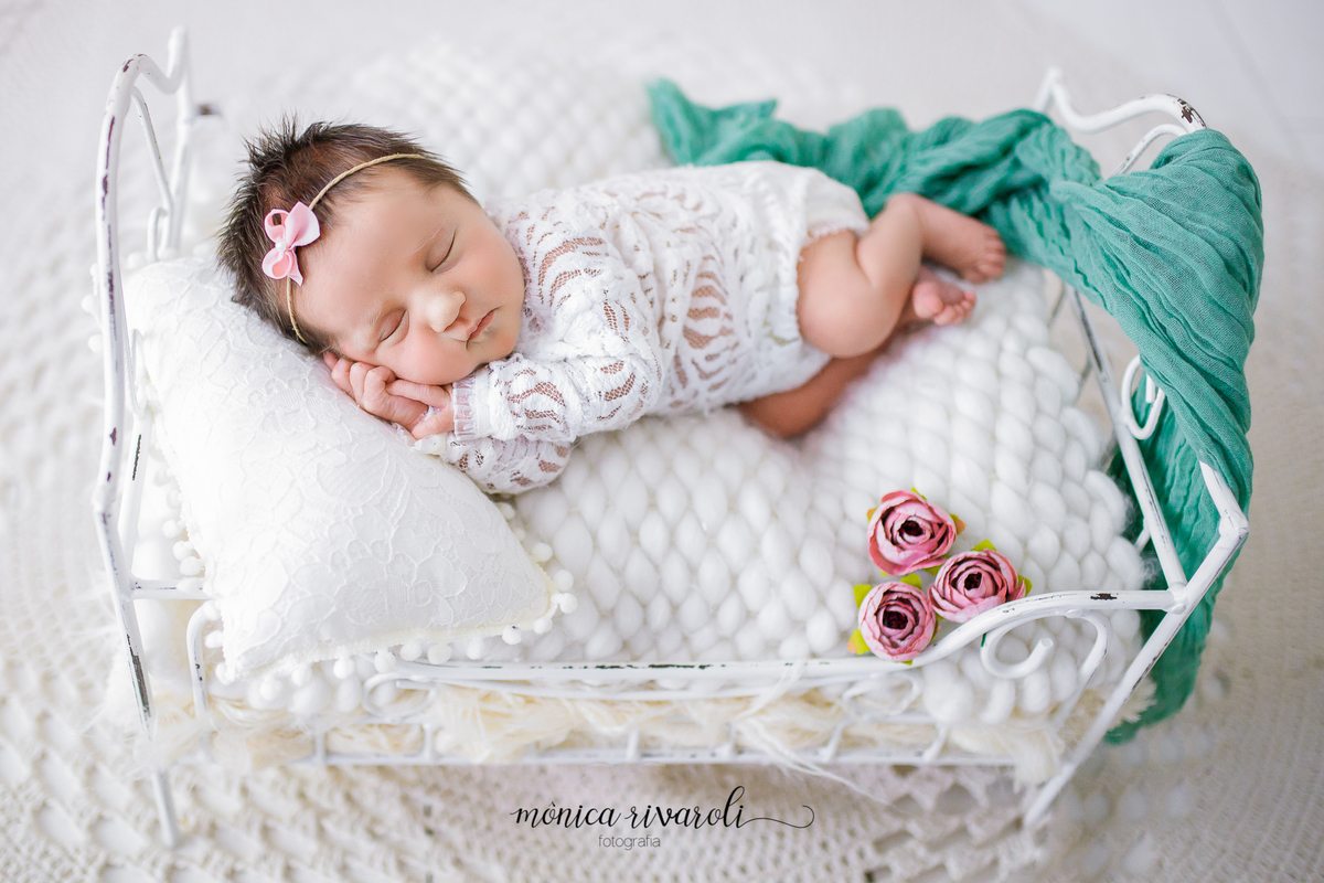 Num fundo branco, a bebê está dormindo em uma caminha de metal branca, com uma manta fofinha e um travesseiro. Ela veste um body de renda também branco e uma tiara com apenas um lacinho rosa. Ela dorme de ladinho, com o rosto apoiado nas mãozinhas. 