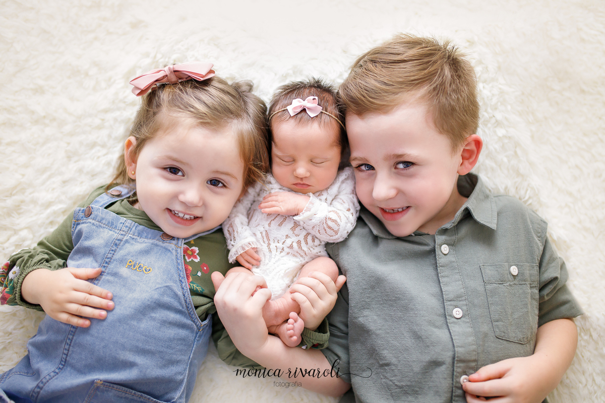 Nesta foto, sobre uma manta de pelinhos, a bebê dorme entre os irmãozinhos, na direita sua irmã e na esquerda o irmão. Os dois tem as mãozinhas na bebê enquanto olham para a câmera e sorriem. 