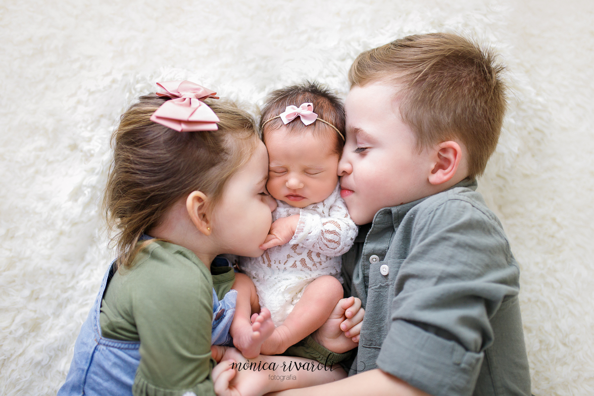 Ainda no mesmo cenário, os irmãos estão deitados de ladinho, cada um de olhos fechados e dando um cheirinho da bebê