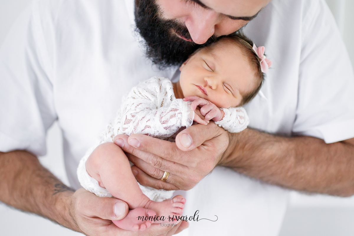 Num fundo branco o papai segura a neném próximo ao seu rosto, enquanto ela dorme nos seus braços, com a mãozinha apoiada no queixo. O papai está de olho fechado e com a cabeça baixa, sobre a neném.