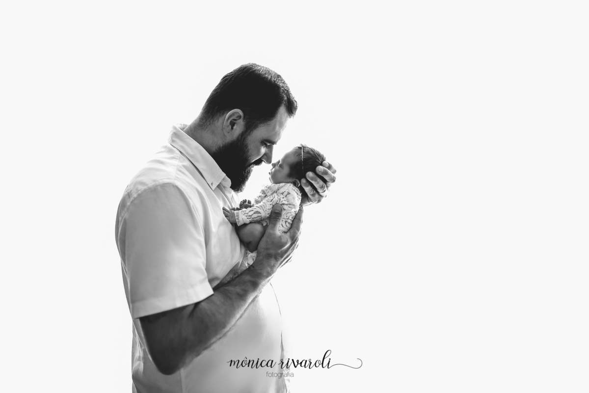 Fundo fundo branco estourado, o papai está de lado com a bebê nas mãos. Com os narizes tocando e os olhos fechados. A foto está em preto e branco. 