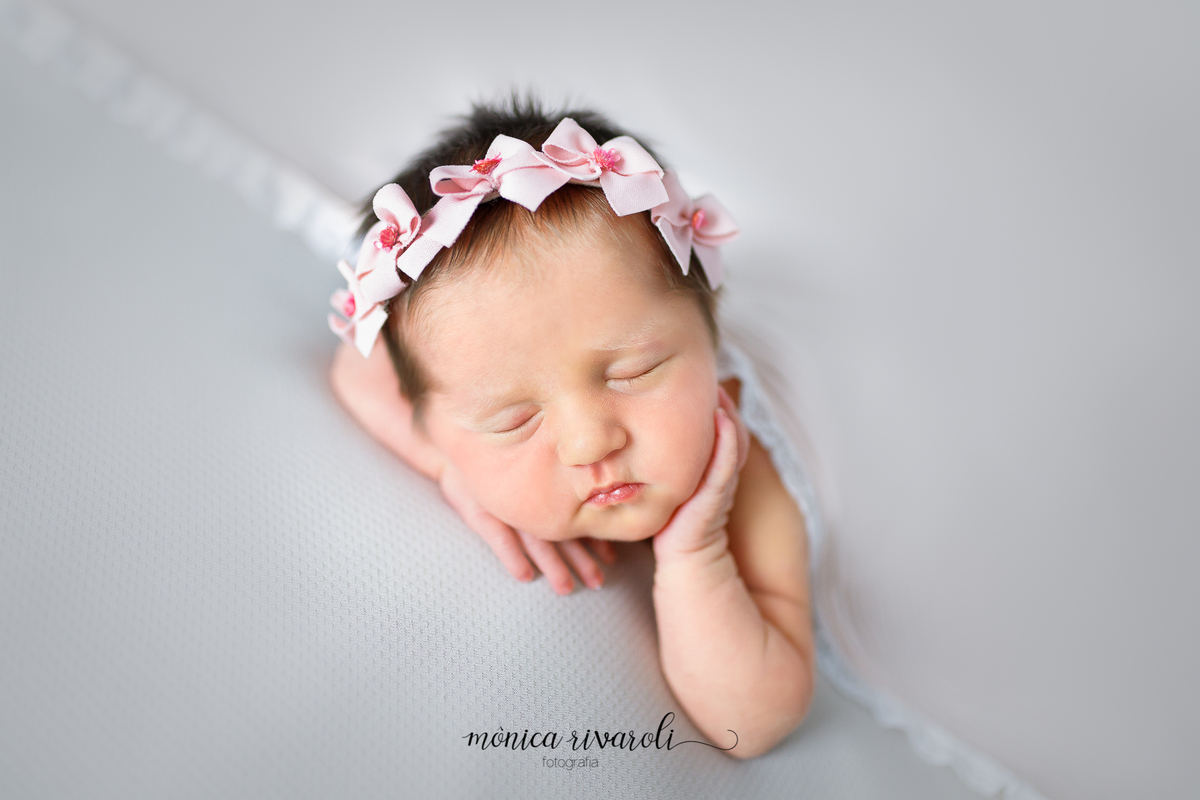 Sobre um tecido cinza, a bebê dorme apoiada em uma das mãozinhas enquanto a outra está na bochecha. Esta coberta por uma manta cinza e tem uma tiara de laços rosas na cabeça. 