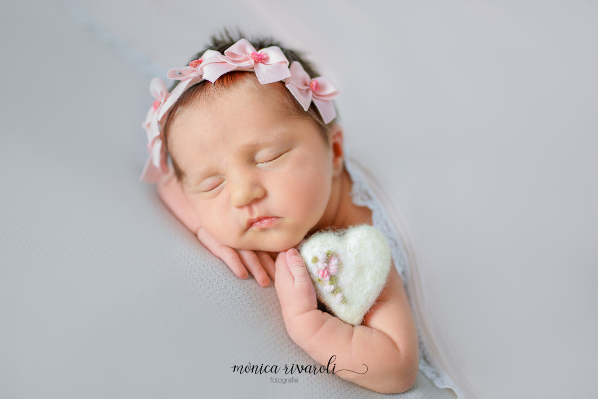 Ainda no tecido cinza e na mesma posição, a bebê agora segura um coração de pelúcia branco e com um ramo de flor rosa, entre o bracinho e o rosto.
