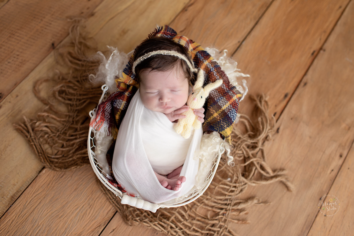Fotografia feita para ensaio Newborn da Betina. Em Studio Localizado em Santo Amaro  - São Paulo, SP. Na foto a bebê está enrolada em um wrep branco. usando uma tiara na cabeça e está dentro de um cesta branca e segura um coelhinho de pelúcia 