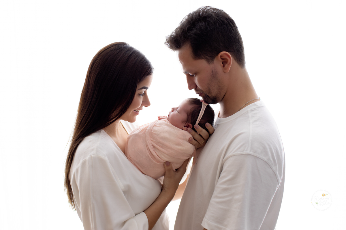 Foto de família feita em ensaio newborn da Betina. Na foto a mamãe e o Papai estão segurando a bebê nas mãos, enquanto encostam as testas. Eles estão de branco e a bebê de rosa. Studio localizado em Jardins - São Paulo, SP.