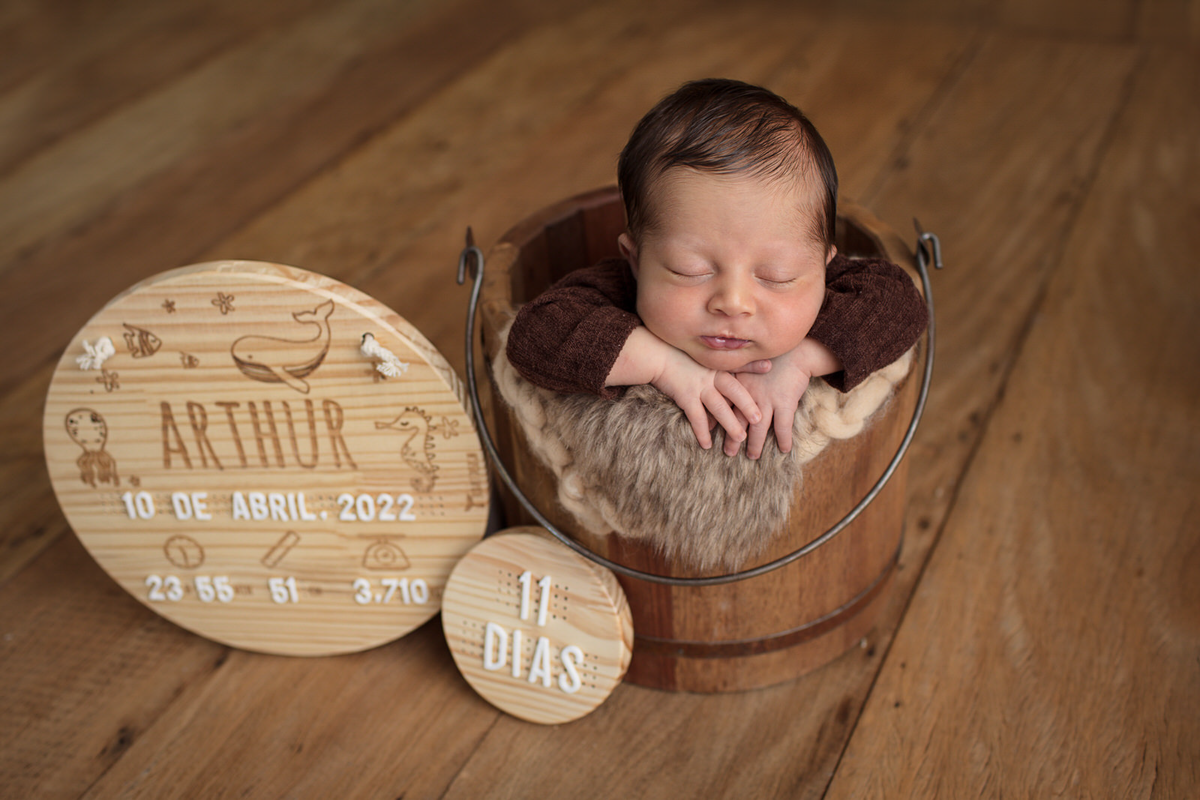 Ensaio newborn do Arthur de 11 dias, em studio localizado na Av. Brigadeiro - São Paulo, SP. Na foto o bebê está dentro de um balde de madeira e ao seu lado um circulo de madeira com seu nome escrito.