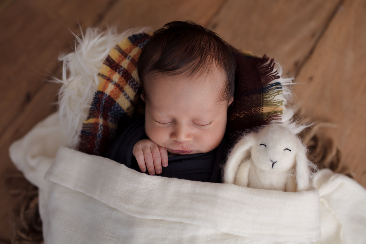 Ensaio newborn do Arthur de 11 dias, em studio localizado em  Moema- São Paulo, SP. Na foto o bebê está deitado em um baldinho de madeira, com tecidos quadriculados, ele está abraçado com um coelhinho branco.