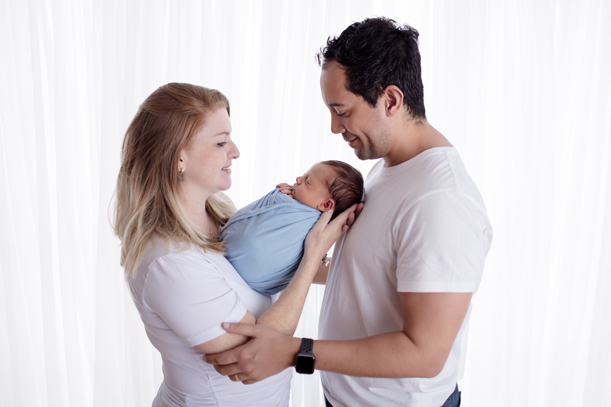 Fotografia em família feita no ensaio newborn do Arthur. Na foto a mamãe está segurando o bebê enquanto o pai olha e sorri de frente para os dois