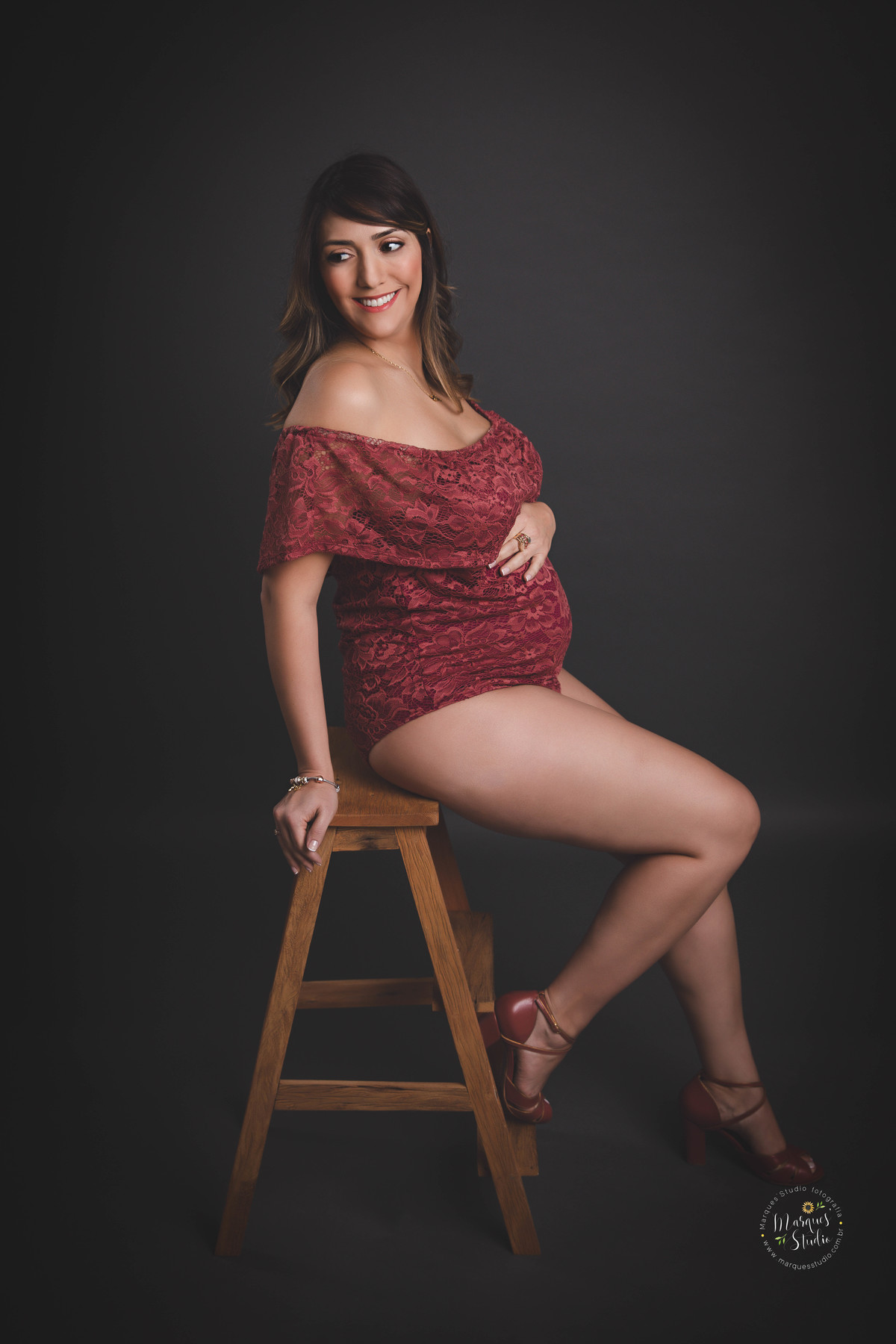 Foto feita para ensaio gestante da Thais, em studio localizado em Osasco - São Paulo, SP. Na foto a mamãe está usando body vinhoe dando um lindo sorriso sentada em um banquinho de madeira.