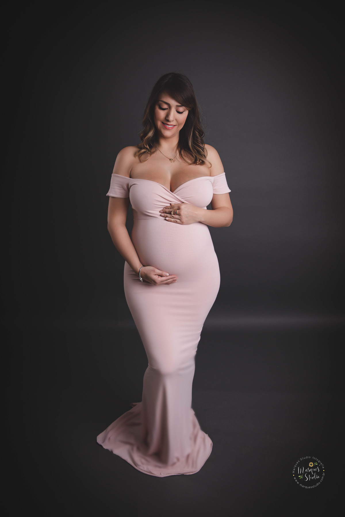 Foto de Studio da gestante Thais, localizado na Avenida Paulista, São Paulo SP, ela está de frente e em pé, com as mãos em sua barriga de gravida, no fundo infinito preto, com um lindo sorriso no rosto, ela usa um lindo vestido rosa