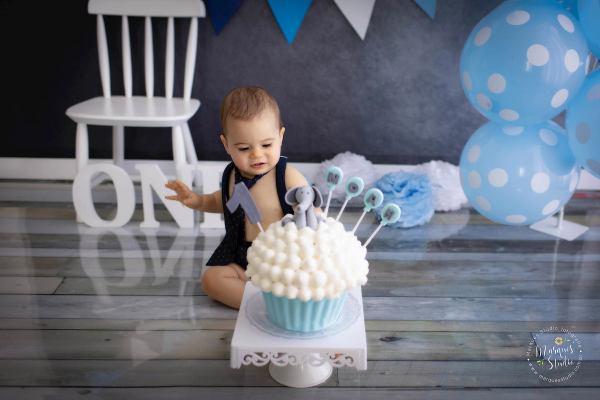 Fotografia de criança de 1 ano em sua sessão de Smash The Cake no studio na Vila Madalena São Paulo SP colocando a mão no bolo branco e azul com a decoração de um elefante cinza e velinha de numero um