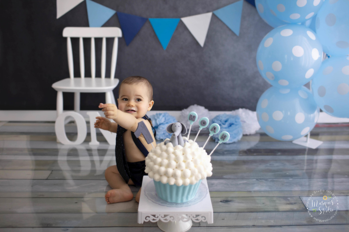 Fotografia de Bebê de 12 meses no ensaio Smash The Cake no studio localizado na Vila Leopoldina São Paulo SP com um lindo bolo nas cores azul e branco com um elefante decorativo com uma cadeira branca balões azuis de bolinhas brancas e bandeiras ao fundo