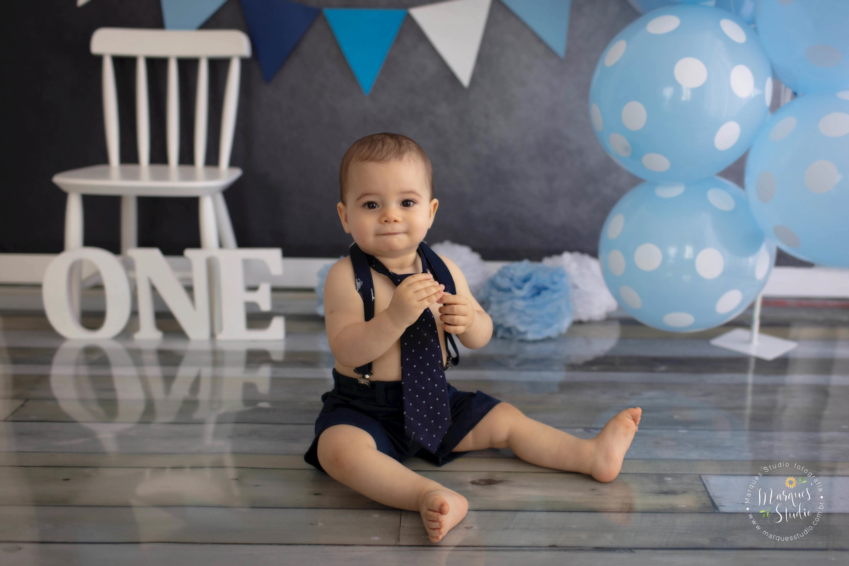 Foto Feita em Studio Fotográfico na zona Oeste Vila Leopoldina São Paulo SP com criança de 12 meses sentada no fundo fotográfico de madeira com decoração de bandeirinhas e balões azuis com uma linda cadeira branca e a palavra one