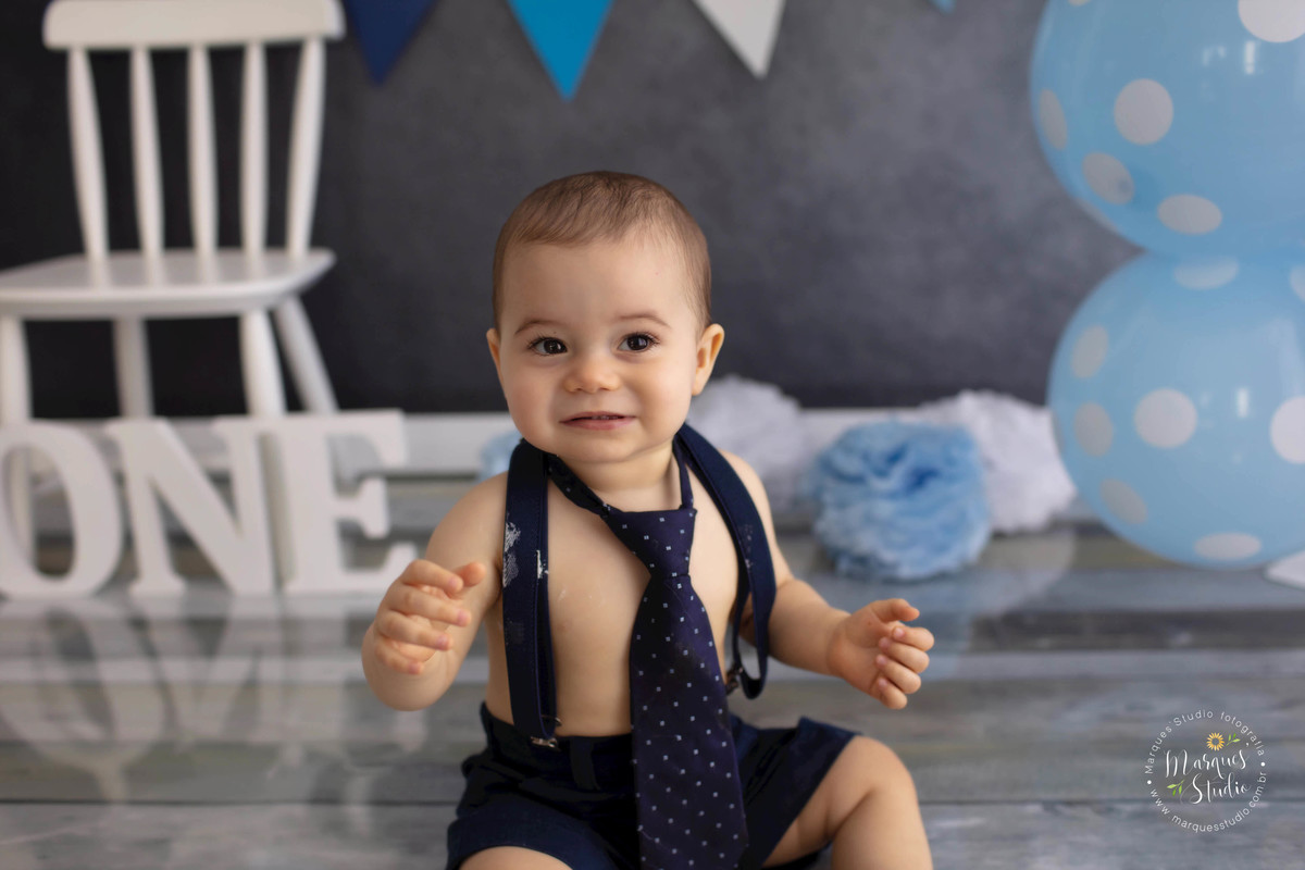 Foto Feita para a sessão Smash The Cake em studio na zona Oeste Osasco SP de criança de 1 ano sorrindo sentada em um fundo fotográfico de madeira com balões e bandeirinhas ao fundo