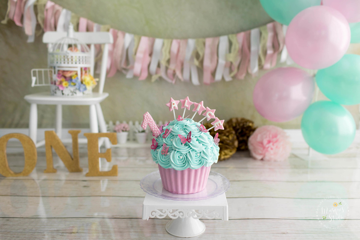 Fotografia de studio com decoração do ensaio smash the cake no fundo fotográfico de madeira com um lindo bolo decorado de borboletas e com uma velinha numero um no centro da foto ao fundo tem balões nas cores rosa e verde bandeirolas coloridas e cadeira