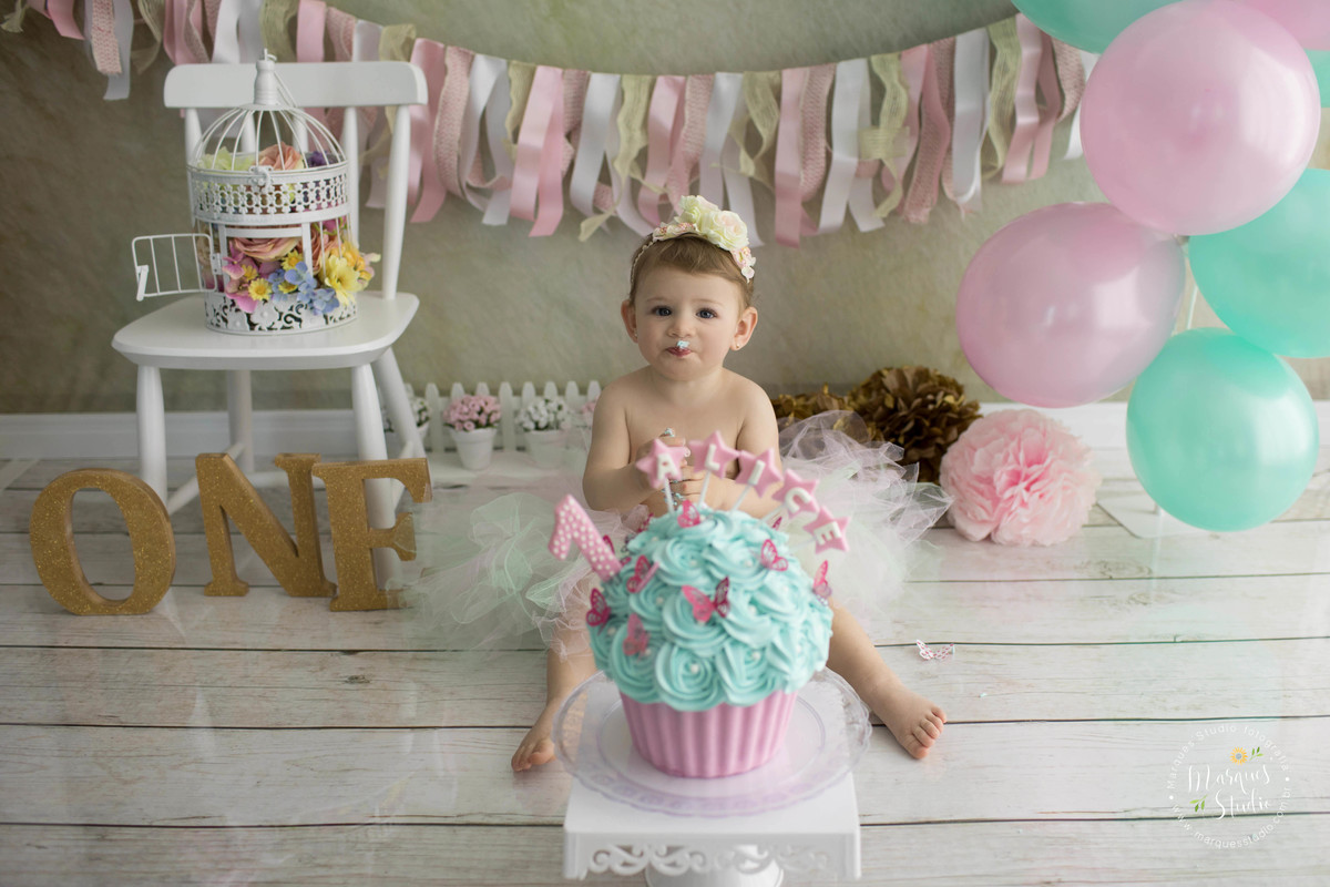 Fotografia feita com menina de 1 aninho para o ensaio smash the cake na Vila Leopoldina SP com balões e bolo rosa com verde Tiffany a bebê está sentada comendo seu delicioso bolo na lateral temos gaiola com flores coloridas