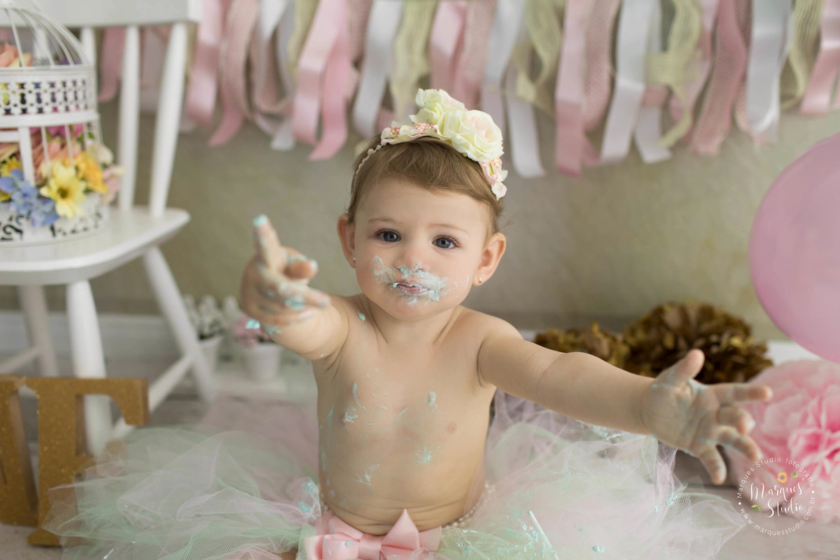 Foto feita em studio fotográfico na zona Oeste São Paulo com uma criança de 1 ano sentada sorrindo e levantando as mãos com bolo ao fundo tem um balão rosa e bandeirolas coloridas uma cadeira branca e uma gaiola com flores dentro