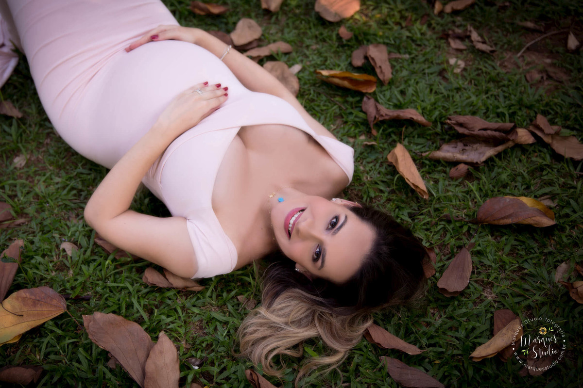 Fotografia feita em um ensaio externo no parque de Osasco São Paulo SP com uma linda gestante que está deitada sobre a linda grama verde com folhas secas ao seu redor ela está com um lindo sorriso no rosto e com as mãos sobre a barriga de gravida 