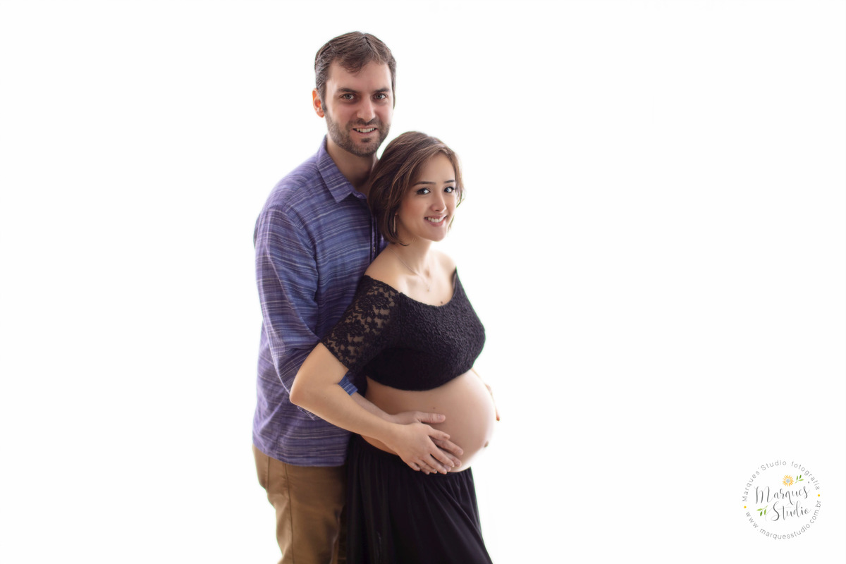 Fotografia de studio fotográfico feito em Osasco São Paulo SP com um casal de grávidos eles estão abraçados ela está na sua frente e ambos estão com as mãos sobre a barriga da gestante com um lindo sorriso ela usa top e saia preto ele usa calca e camisa