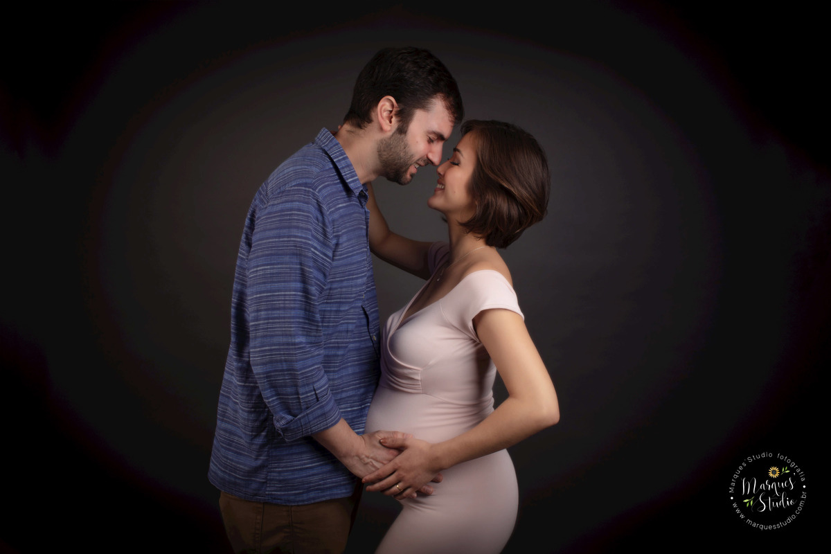 Fotografia feita no studio de Osasco São Paulo SP com um lindo casal de grávidos eles estão um de frente para o outro colocando as mãos sobre a barriga da gestante ela usa um lindo vestido longo rosa e ele calca e camisa estão no fundo infinito preto