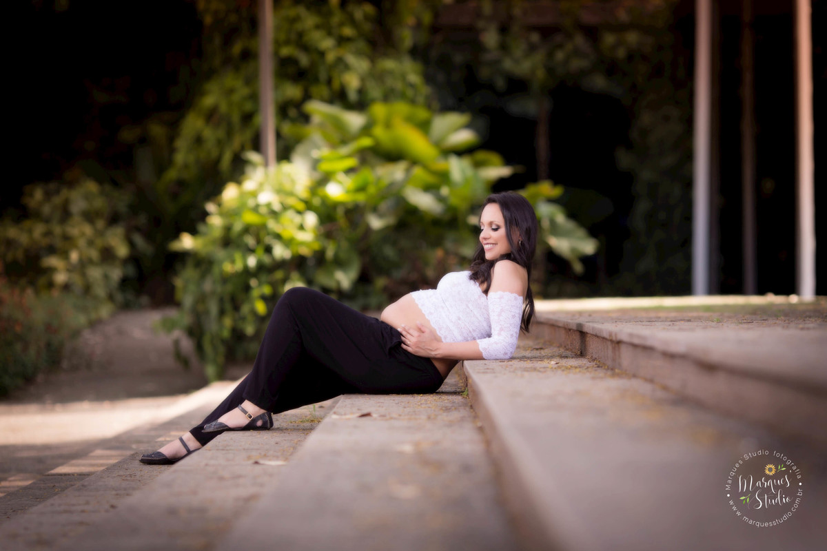 Fotografia realizada em um parque de São Paulo, SP, Morumbi, para um ensaio de gestante com 32 semanas de gestação, Eliane está sentada em uma escada olhando para sua barriga de gravida, ela usa um lindo top branco em renda, com uma linda saia preta
