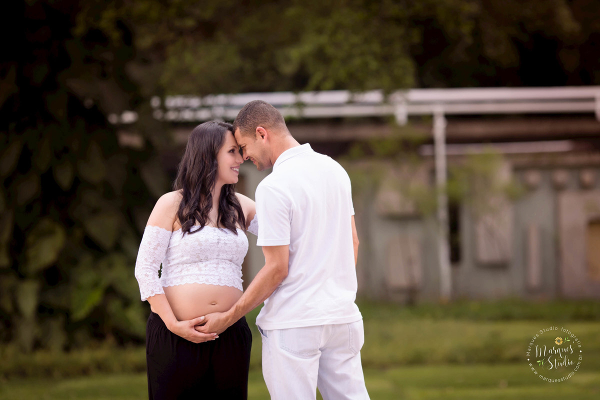 Foto feita no ensaio externo da gestante Eliane, feito em Perdizes São Paulo, SP, na fotografia temos um casal de grávidos sorrindo um para o outro colocando as mãos sobre a barriga da gravida, ao fundo da foto uma linda arvore com muitas flores