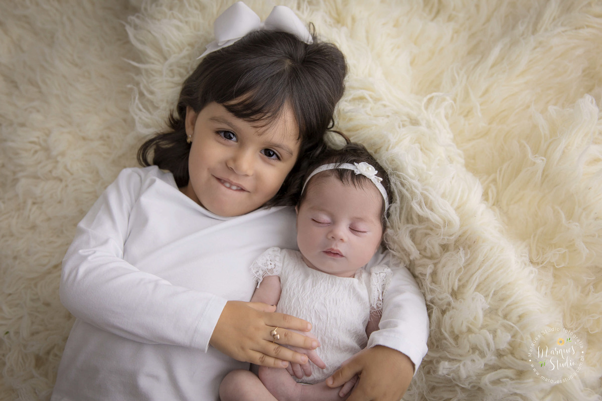 Foto de ensaio newborn,  recém-nascida abraçada com a irmã em ensaio fotográfico de bebê na vila Madalena em São Paulo, SP. As irmãs usam roupinhas na cor branca, com um lindo laço na cabeça.