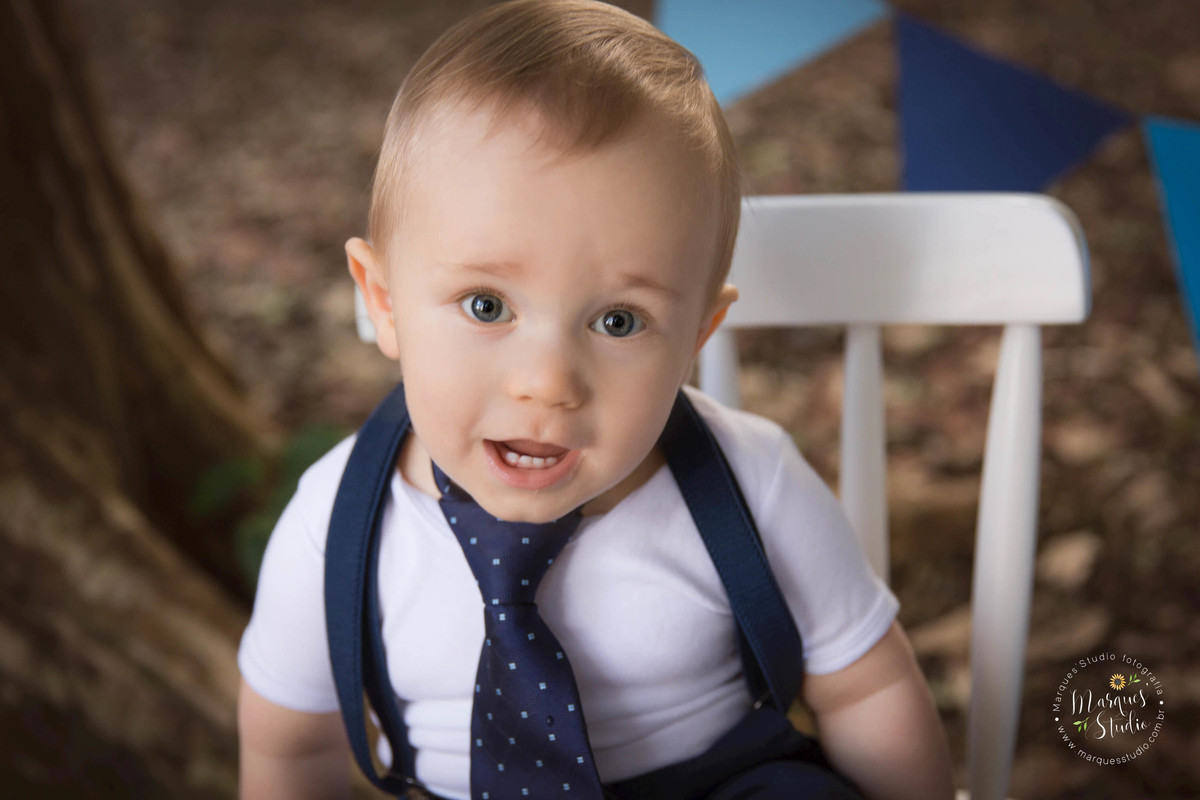 Foto Feita para a sessão Smash The Cake externa, na zona Oeste, Osasco, SP, de uma criança de 1 ano sorrindo sentada em uma cadeirinha branca, com balões e bandeirinhas ao fundo, ele está com um conjunto de short, camisa, gravata e suspensório.