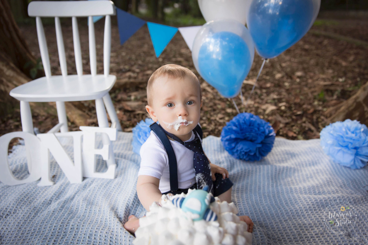 Fotografia de Bebê sentado comendo um bolo em sua sessão de  Smash The Cake no parque em Osasco São Paulo SP, sua mãozinha direita está no bolo, ao fundo temos uma linda decoração na cor azul e branco, com balões e bandeirolas, cadeira e folhagem.