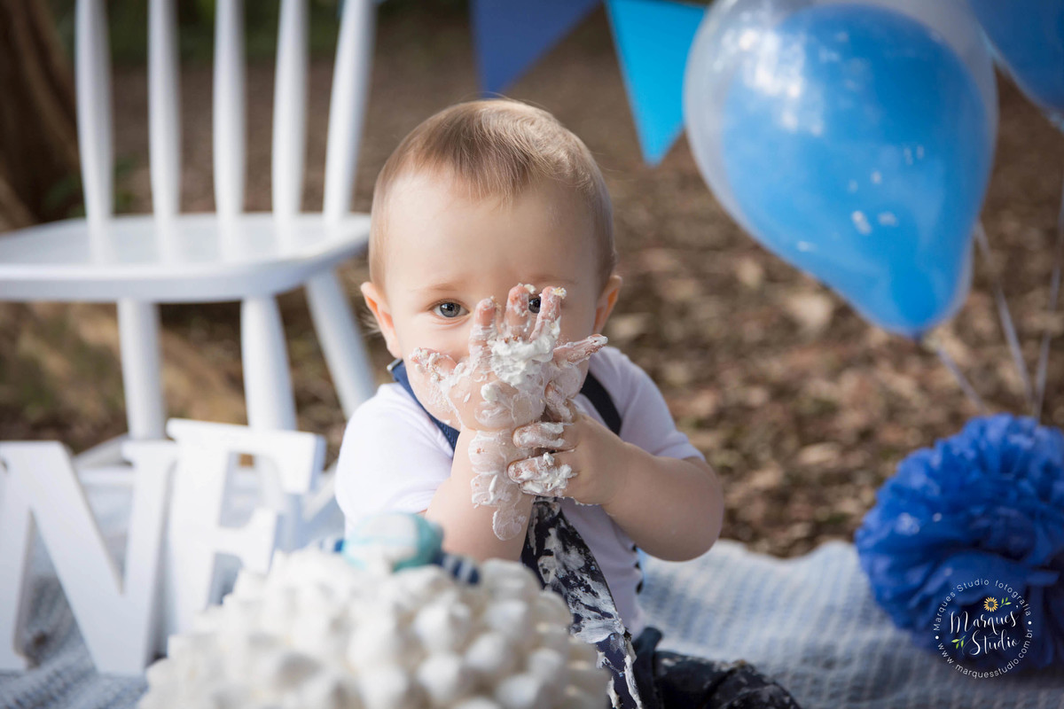 Fotografia de Bebê de 12 meses no ensaio Smash The Cake no parque localizado na Vila Leopoldina São Paulo SP com um lindo bolo nas cores azul e branco, uma cadeira branca balões azuis, e bandeiras ao fundo, o bebê está sentado e comendo o delicioso bolo.