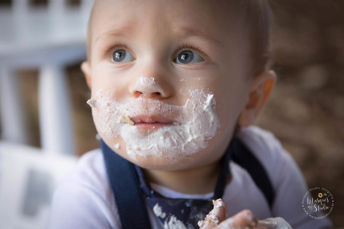 Foto do rosto de uma criança de doze meses, em sua sessão de smash the cake, para o seu acompanhamento, ele está com bolo em seu rosto, e está usando um lindo conjunto azul e branco, seus olhos são azuis, ele está no parque na Avenida Paulista, São Paulo.
