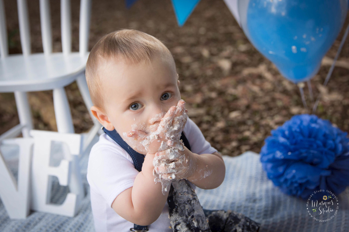 Fotografia de bebê de 1 ano comendo um delicioso bolo no ensaio Smash The Cake no parque na Avenida Paulista, São Paulo, SP, o bebê está com bolo em sua mão e em sua boca, ao fundo uma linda decoração nas cores azul e branco.