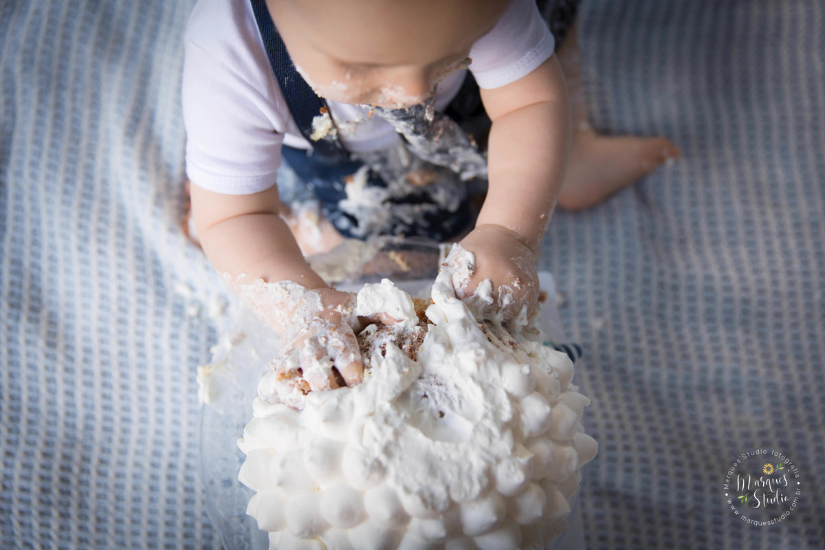 Fotografia de uma sessão smash the cake, realizada na Vila Perdizes, São Paulo, SP, na foto temos um menino que amassa o bolo com suas lindas mãozinhas, ele está usando um lindo conjunto de short azul e camiseta branca, e suspensório azul marinho. 