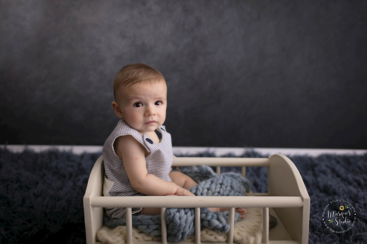 Foto feita em studio fotográfico localizado na Vila Madalena, São Paulo, SP, com uma criança de 6 meses sentada em uma caminha de madeira, ele está sorrindo, o fundo fotográfico é na cor azul marinho, o bebê está usando um lindo macacão azul.
