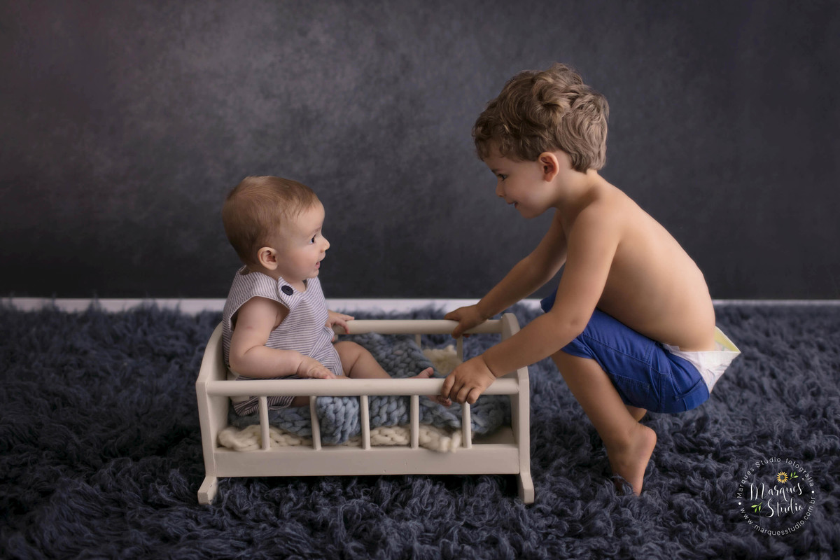 Fotografia de ensaio em studio fotográfico localizado na Vila Perdizes, São Paulo, SP, com uma criança de 6 meses sentada em uma caminha de madeira, ele está sorrindo para o irmãozinho que está de frente pra ele, o fundo fotográfico é na cor azul marinho.