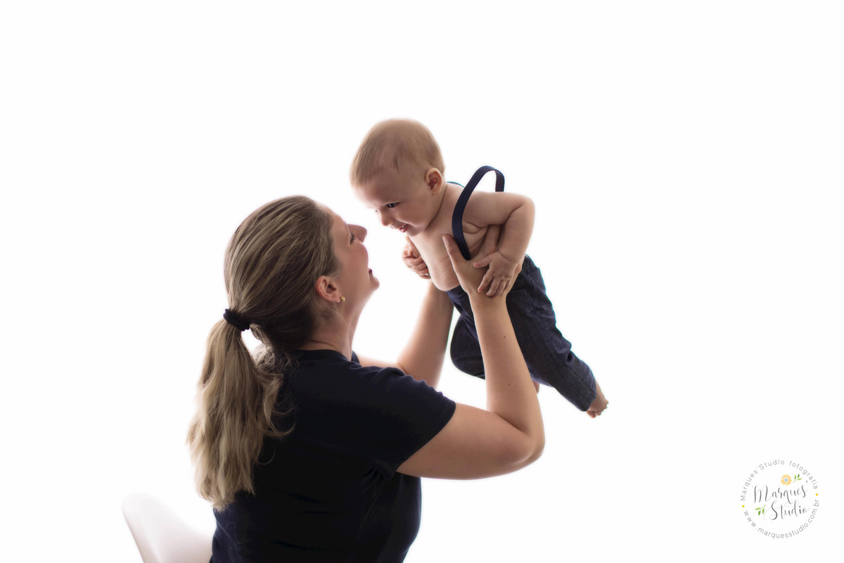 Foto da sessão de acompanhamento de 6 meses do Leo, no  Studio, na Vila Madalena, São Paulo, SP, na foto temos a mamãe de costas segurando o bebê no colo que está dando um lindo sorriso, eles estão no fundo fotográfico branco.