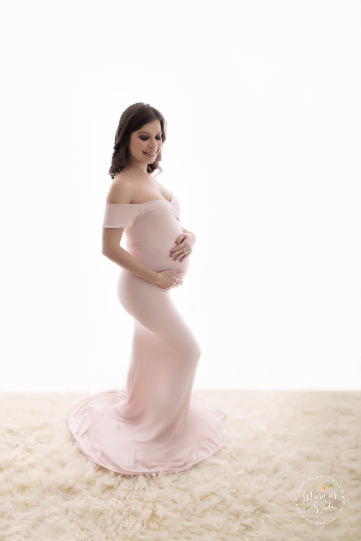 Fotografia em Studio da gestante Clara, localizado na Vila Perdizes São Paulo SP, ela está de perfil e em pé, com as mãos em sua barriga de gravida, no fundo infinito branco com um lindo sorriso no rosto ela usa um belíssimo vestido longo na cor rose.