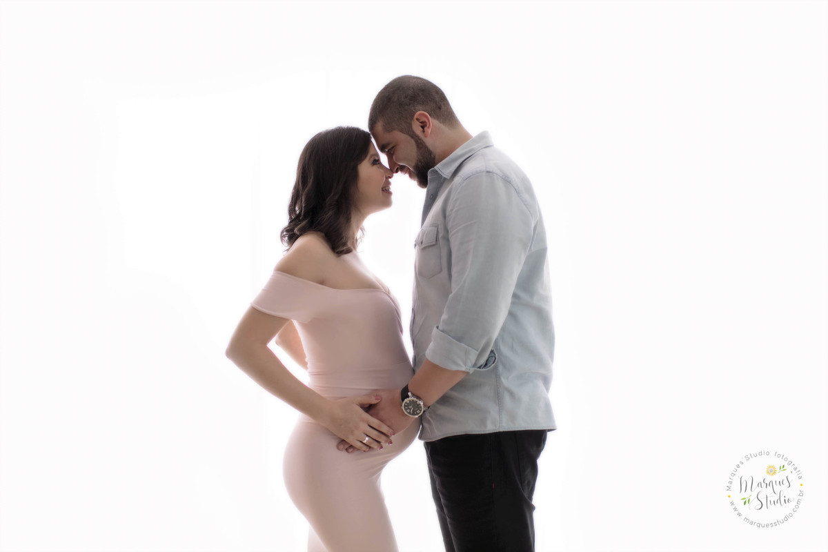 Foto feita para ensaio gestante casal se olhando com as mãos na barriga da gravida ela usando um lindo vestido longo rosa e seu esposo usando uma camisa jeans calça jeans preta, os dois com um lindo sorriso no fundo infinito branco, no studio em Osasco SP