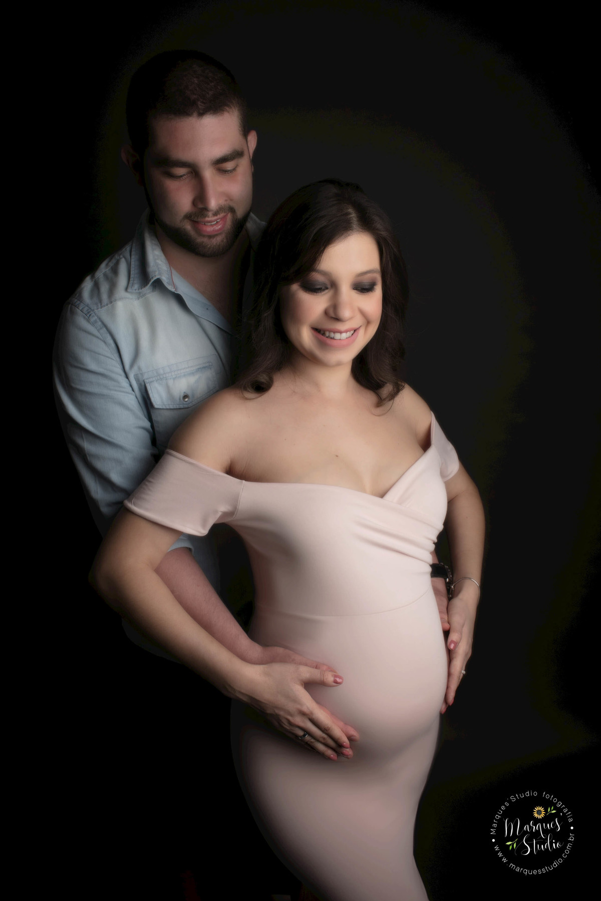 Fotografia de um casal de grávidos, a mamãe está na frente do papai, os dois estão com as mãos sobre a barriga da gestante, eles estão sorrindo, ela usa um lindo vestido longo rosa, e ele usa uma camisa e calça jeans, eles estão no studio em Pinheiros, SP