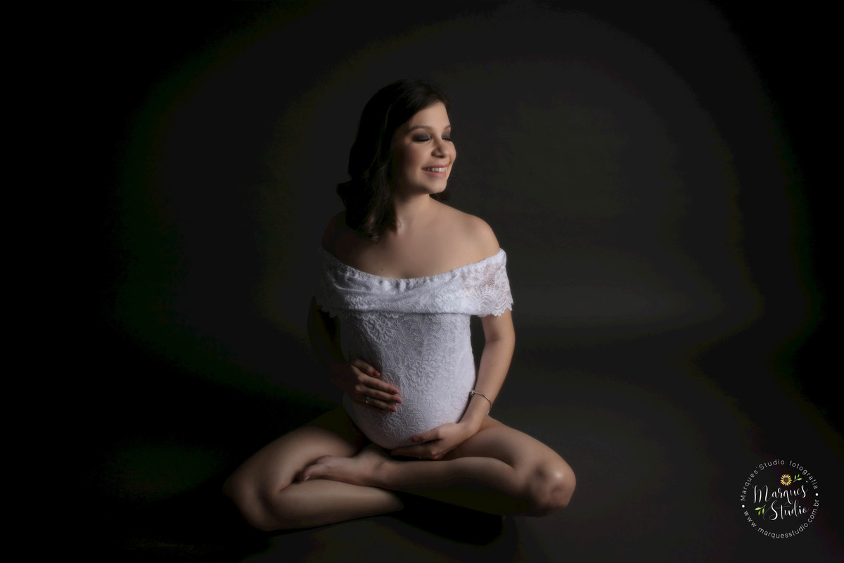 Foto de gestante em Studio na Vila Leopoldina, São Paulo, SP a mamãe está sentada de perna cruzada de índio, ela está com as mãos sobre a sua barriga gravida com um belo sorriso estampado no rosto, ela está usando um body branco com renda,  no fundo preto