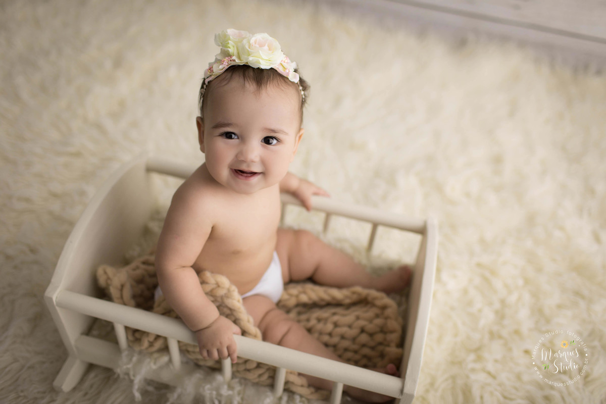 Fotografia de uma linda bebê para a sua sessão de acompanhamento infantil de 6 meses, ela está sentada em uma caminha de madeira, e está dando um belo sorriso, ela usa uma linda headband na cor bege com flores rosas, o estúdio fica na Vila Leopoldina, SP.
