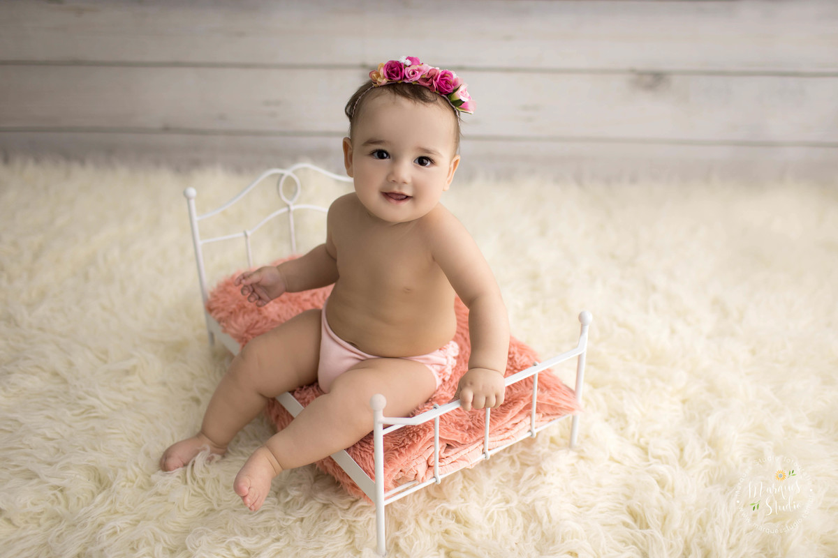 Fotografia de bebê menina para a sua sessão de acompanhamento infantil de 6 meses, ela está sentada em uma caminha de ferro, e está dando um belo sorriso, ela usa uma linda headband na cor rosa com lindas flores, o estúdio fica no Morumbi, São Paulo, SP.