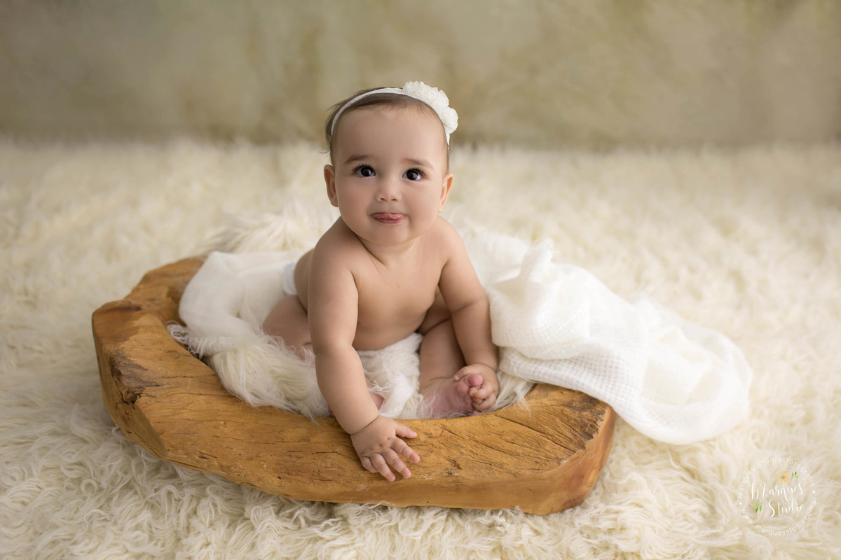 Foto feita em studio fotográfico localizado na zona Oeste São Paulo, SP, com uma criança de 6 meses sentada em um cesto, ela está sorrindo, com as mãozinhas no pezinho, ela está em um lindo fundo fotográfico na cor bege, com um lindo tapete bege.