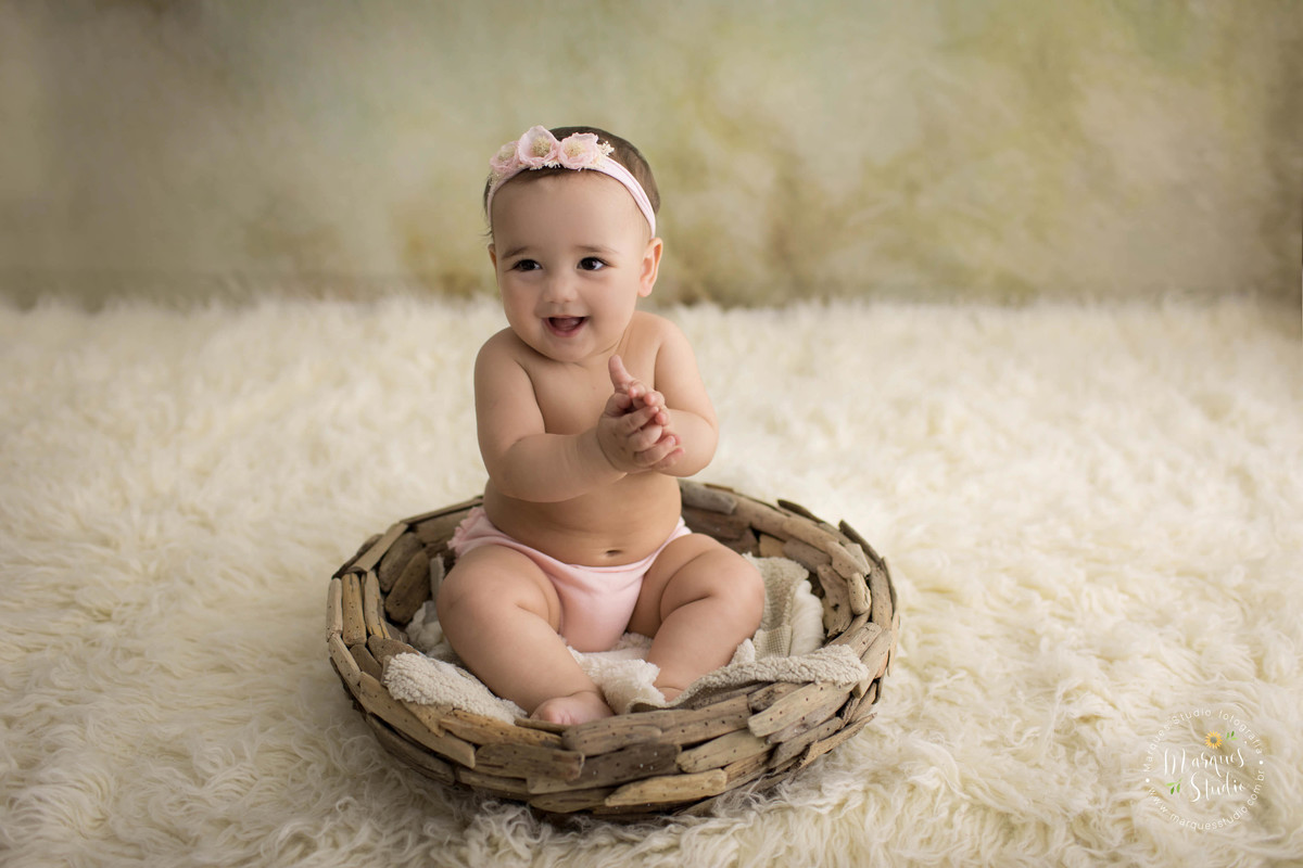 Foto feita em studio fotográfico localizado em Pinheiros, São Paulo, SP, com uma criança de 6 meses sentada em um cesto, ela está sorrindo, batendo palma, ela está em um lindo fundo fotográfico na cor bege, com um tapete bege, ela usa um laço de flores.
