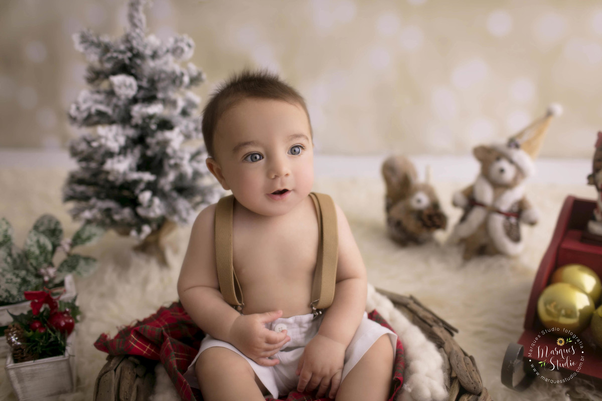 Fotografia para o ensaio de Natal do Enrico, em nosso studio localizado na Vila Perdizes, São Paulo, SP, o bebê está sentado em um cesto de madeira, ao seu redor tem arvore de Natal o Papai Noel e um urso, o bebê de 6 meses está dando um lindo sorriso.