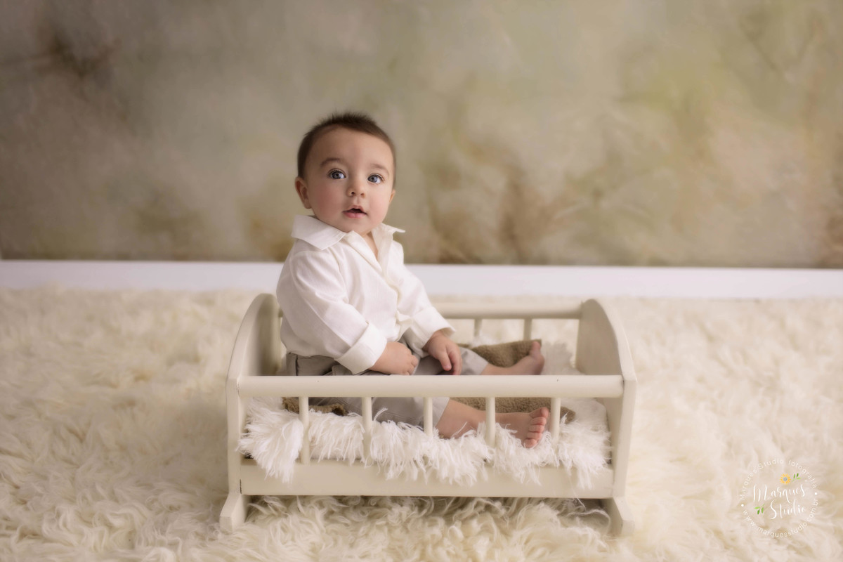 Foto feita em studio fotográfico localizado na Vila Madalena, São Paulo, SP, com uma criança de 6 meses sentada em uma caminha de madeira, ele está sorrindo, e está no fundo fotográfico bege, com um tapete na cor bege, ele usa conjunto de calça e camisa.