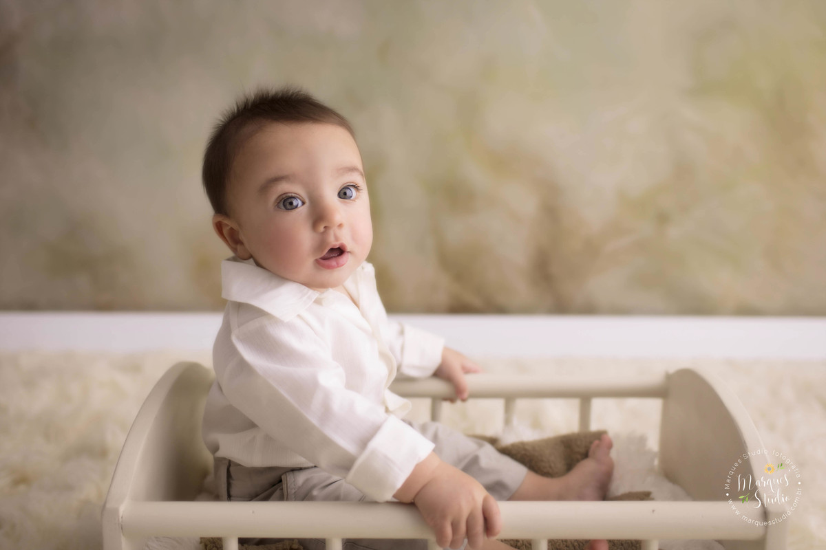 Fotografia para o book de bebê do Enrico, no studio fotográfico na Vila Leopoldina, São Paulo, SP, na foto o bebê está sentado de perfil em uma cama de madeira, no fundo fotográfico bege, ele está com as mãos na caminha e usando conjunto de calça e camisa
