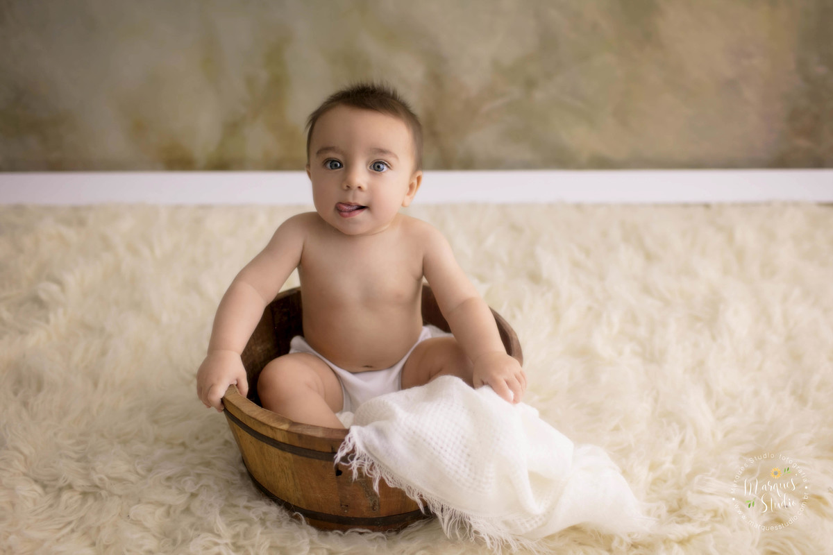 Foto feita em studio fotográfico localizado na zona Oeste, Osasco, São Paulo, SP, com uma criança de 6 meses sentada em um cesto de madeira, ele está sorrindo, o fundo fotográfico é na cor bege e branco. 