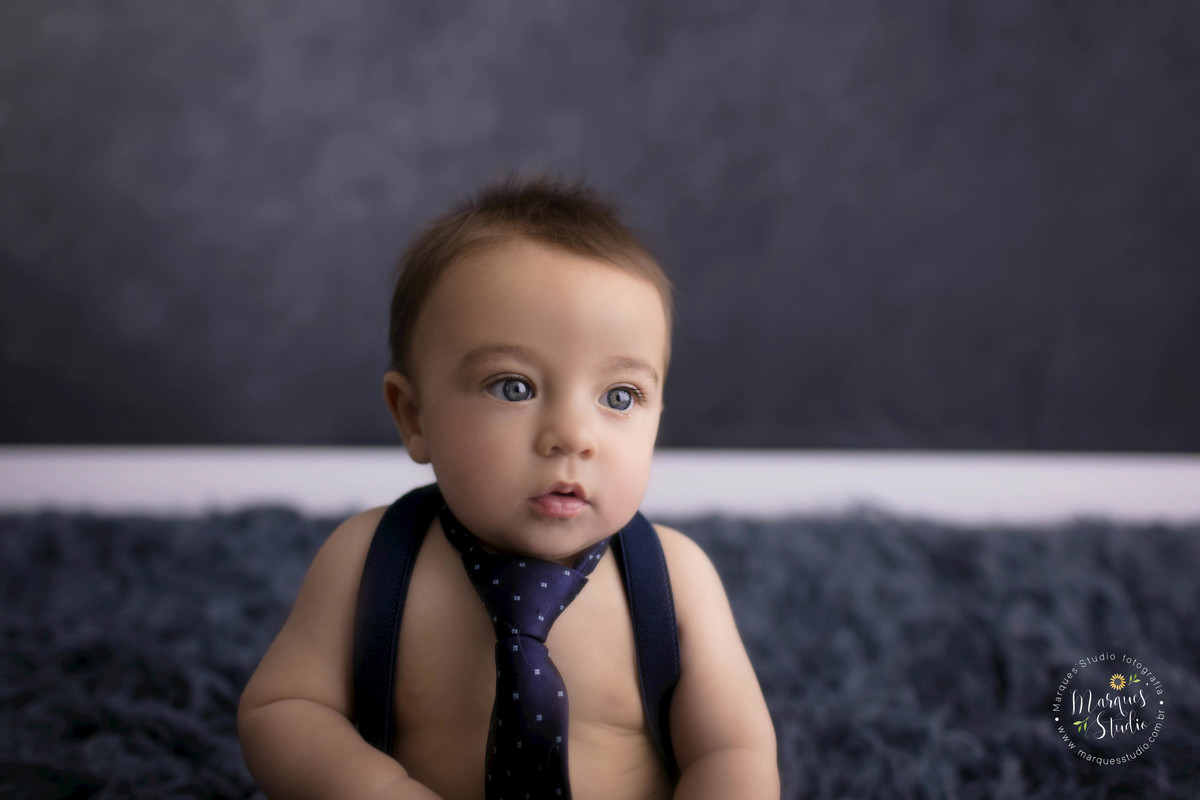 Foto Feita para a sessão de acompanhamento infantil em studio na zona Oeste, Sumaré, São Paulo, SP, de uma criança de 6 meses sorrindo, ele está sentado em um lindo tapete azul marinho, e o fundo fotográfico é na cor azul marinho, ele está de gravata.