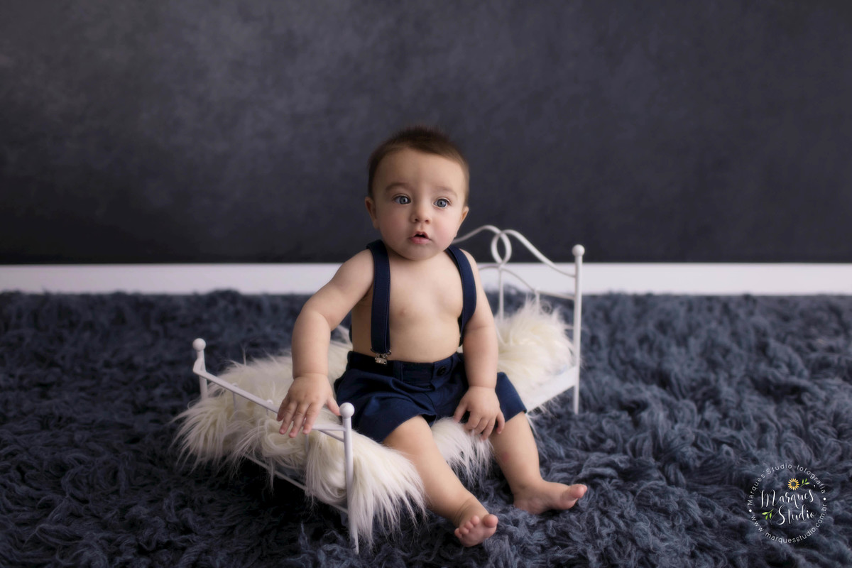 Foto feita em studio fotográfico localizado na Vila Madalena, São Paulo, SP, com um bebê de 6 meses sentado em uma caminha de ferro, ele está sorrindo, o fundo fotográfico é na cor azul marinho, o bebê está usando um lindo conjunto de short e suspensório.